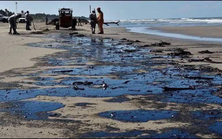playa con petróleo - PUNTO DE ENCUENTRO Una fotografía de la playa que captura un esfuerzo de limpieza tras un derrame-de-petróleo. El suelo está cubierto de charcos oscuros y viscosos de petróleo, mientras que el mar se extiende hacia el horizonte bajo un cielo soleado.