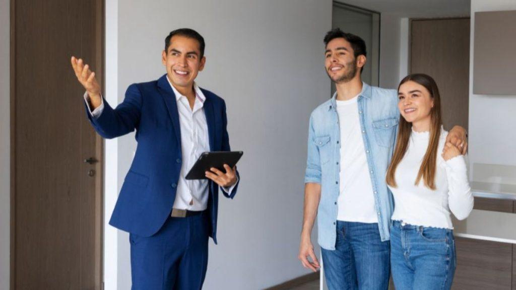 Real estate agent in a blue suit gestures as he explains a property to a smiling couple in a modern interior, holding a tablet.