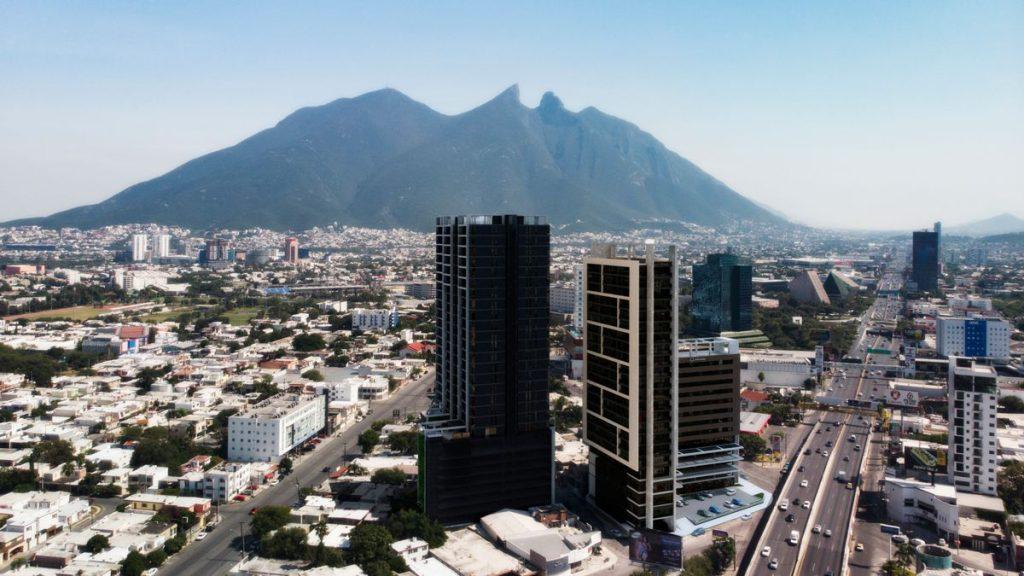 Aerial view of a city with two tall dark skyscrapers and a wide road, mountains in the distance under a blue sky.