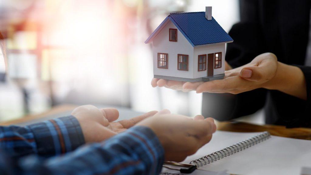 desarrollador-inmobiliario-casa-monterrey-quantium-desarrollos - PUNTO DE ENCUENTRO Hands presenting a small model house to someone across a desk, symbolizing real estate or home ownership.