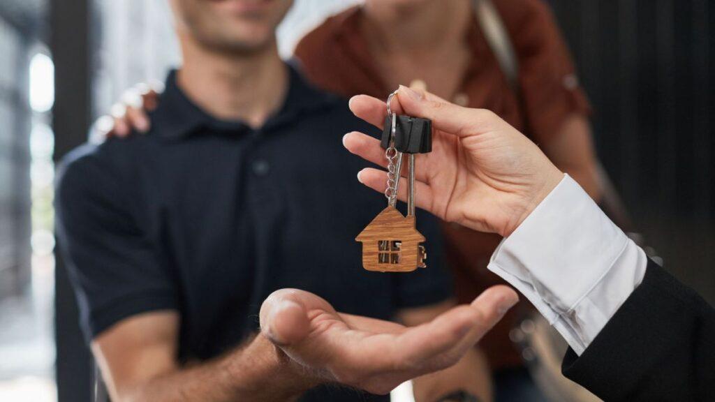 comprar-un-inmueble-resistente-a-cortes-de-luz - PUNTO DE ENCUENTRO Close-up of a wooden house keychain being handed over to a recipient, symbolizing a home purchase or rental transfer