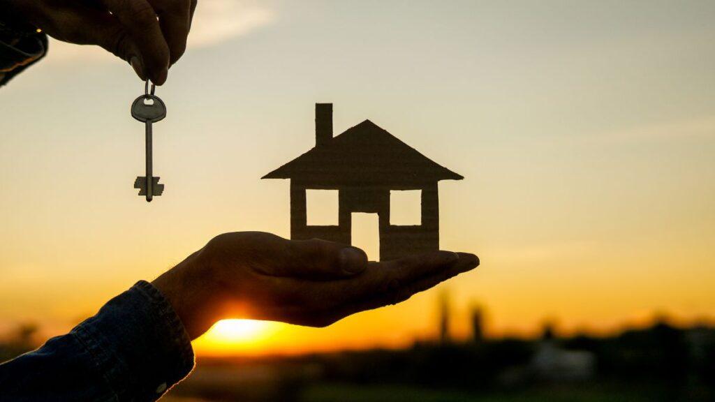 cuánto-vale-mi-casa-quantium-desarrollos-avalúo-inmobiliario - PUNTO DE ENCUENTRO Person holds a key above a hand-shaped cutout of a house at sunset, symbolizing home ownership or real estate.