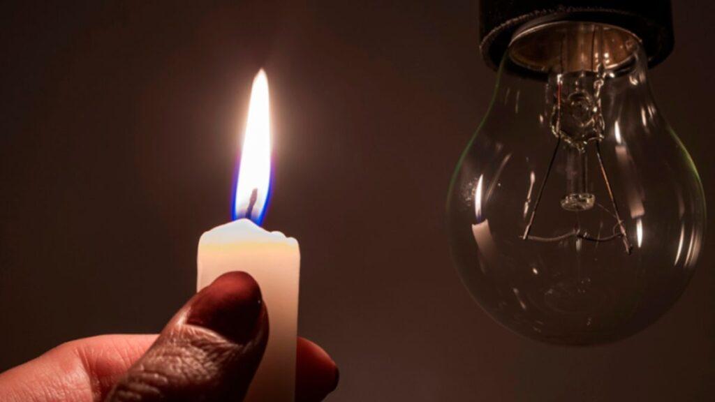que-revisar-antes-de-comprar-una-casa-en-monterrey - PUNTO DE ENCUENTRO Hand holding a lit white candle beside a clear incandescent light bulb in a dark room, contrasting flame and bulb.