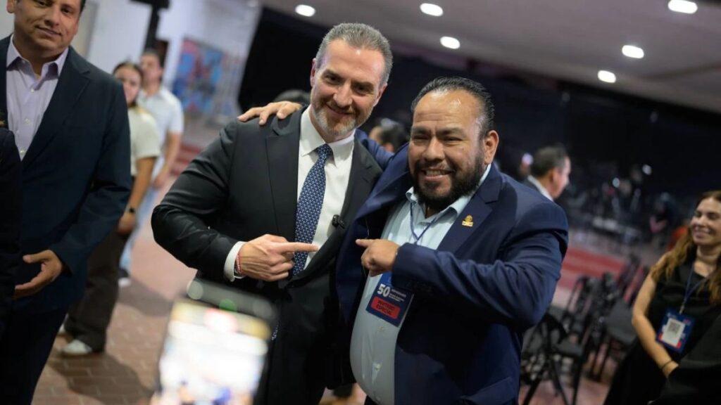 Two men in business suits smile for a photo at a conference, one pointing at the other.