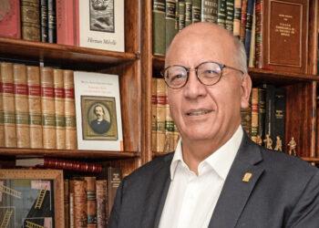 Smiling older man in glasses and a dark suit stands in a library with tall wooden bookshelves behind him.”,