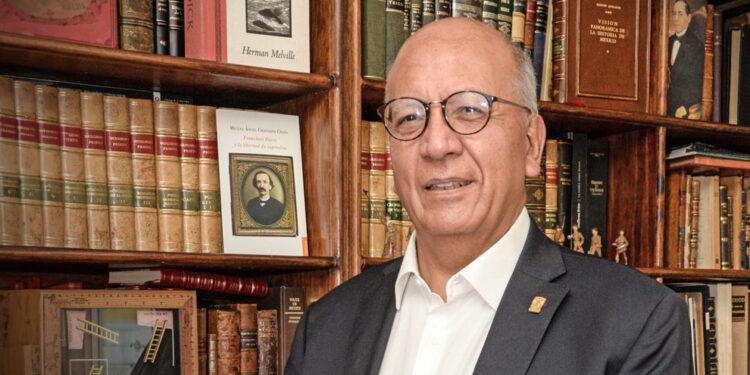 vicente-quirarte - PUNTO DE ENCUENTRO Smiling older man in glasses and a dark suit stands in a library with tall wooden bookshelves behind him.”,